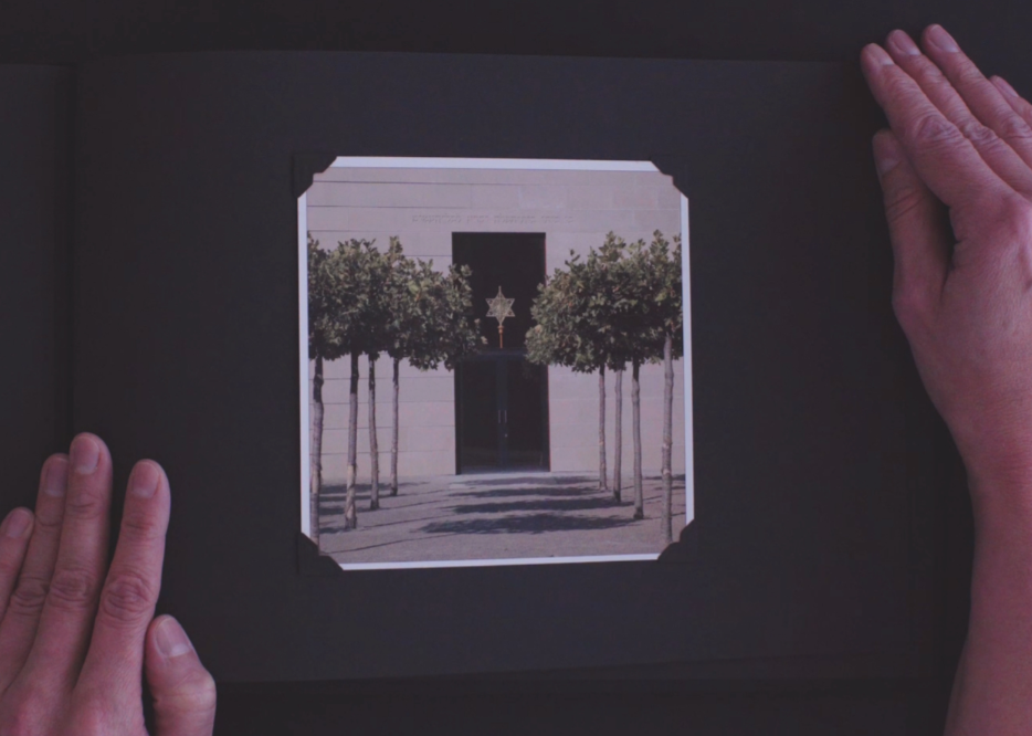 Filmstill: Hände halten ein Fotoalbum mit einer historischen Aufnahme einer Synagoge